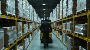 Une image dans un entrepôt de reverse logistique avec un opérateur qui manœuvre des palettes au milieu d'étagères remplies de produits de retour colis.